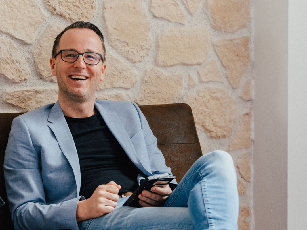Ein Immobilienmediator sitzt entspannt in einem Sessel vor einer hellen Steinwand und lächelt in einer modernen Innenumgebung.