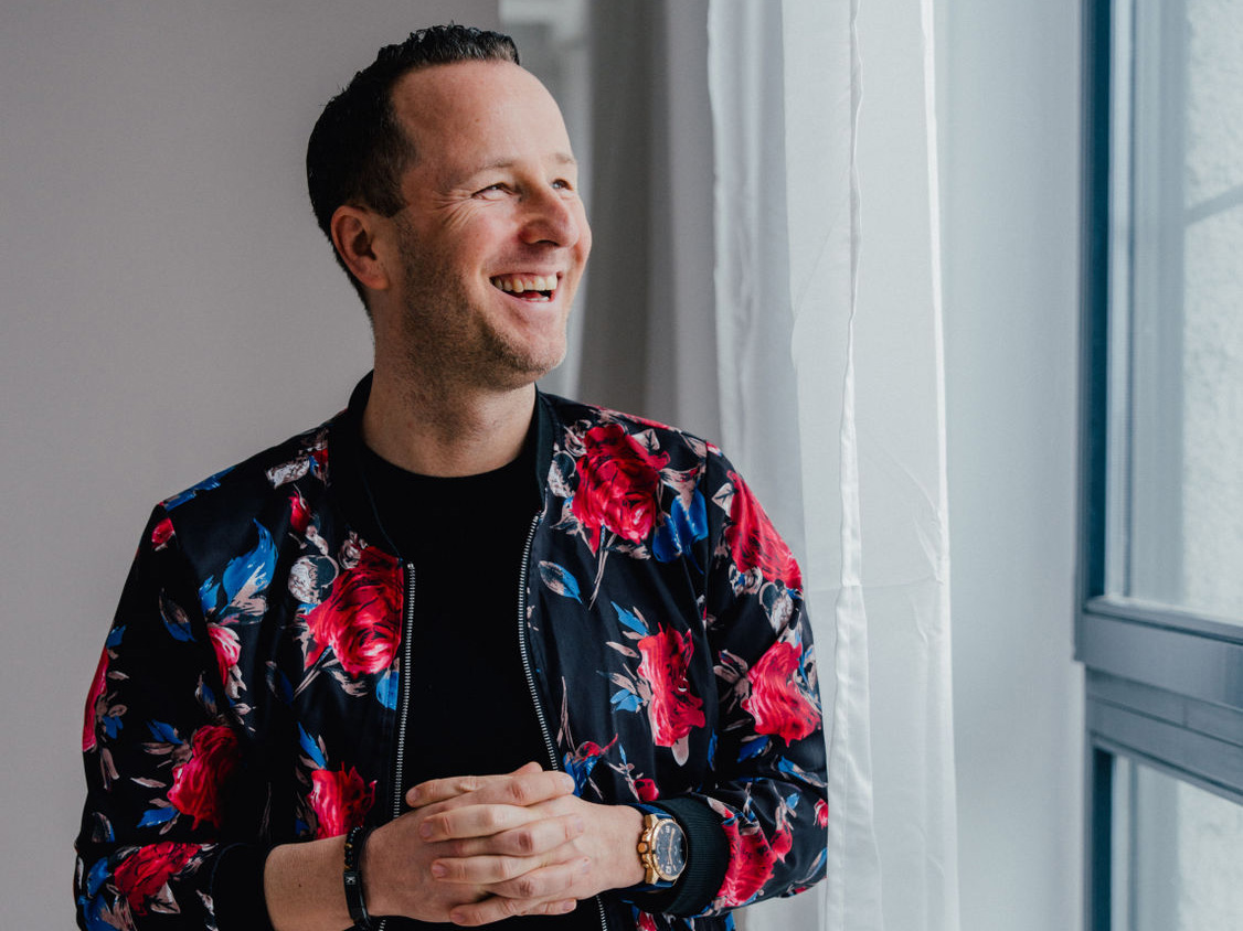 Stefan Pfaff steht lächelnd vor einem Fenster und trägt ein gemustertes Sakko in einer hellen Innenumgebung.